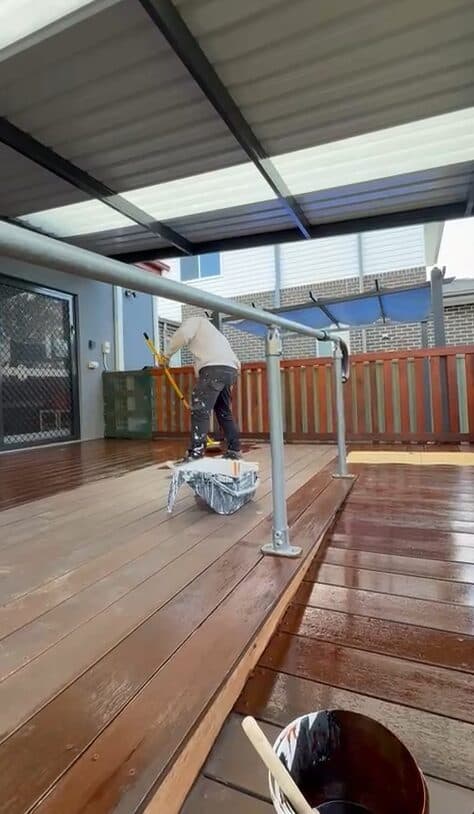 Before — VD Painting crew member applying stain to a weathered timber deck under a patio in NSW