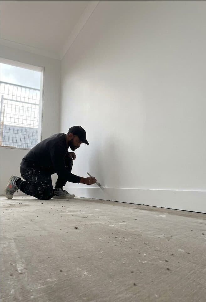 Before — VD Painting crew member cutting in fresh white paint along the skirting board of a VIC interior wall (work in progress)